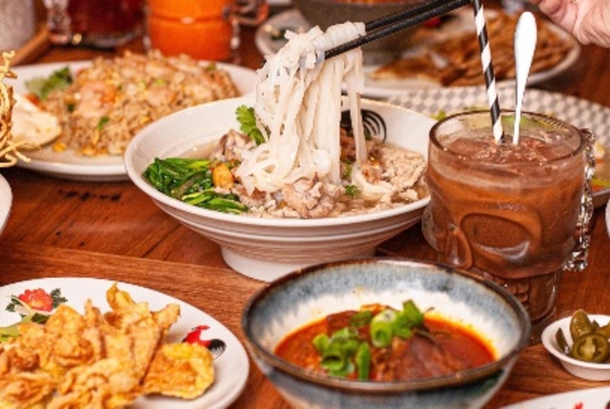 Malaysian dishes of soupy noodles, fried rice, a curry, and an iced coffee in a glass, on a cafe table top.