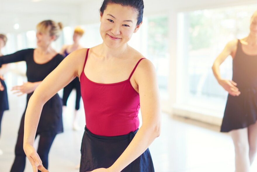 A woman in a red leotard and black skirt practicing a ballet position in a lightfilled ballet studio with other dancers in the background.