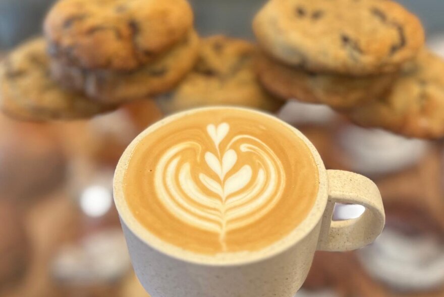 A flat white coffee in a cup with milk froth art, behind are slightly out of focus choc-chip cookies.