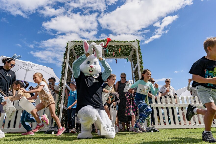 Children running past a person in an Easter bunny costume and a white picket fence during an outdoor easter egg hunt under a blue sky. 