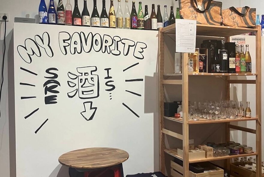 A sign with an arrow pointing to an empty stool next to a bookshelf full of glasses and bottles, sign reading 'my favourite sake'.