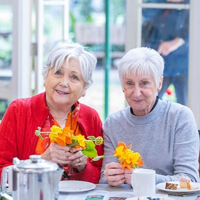 Two older women seated with crafts, tea and cakes in an indoor setting.