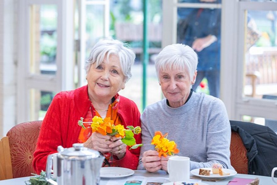 Two older women seated with crafts, tea and cakes in an indoor setting.