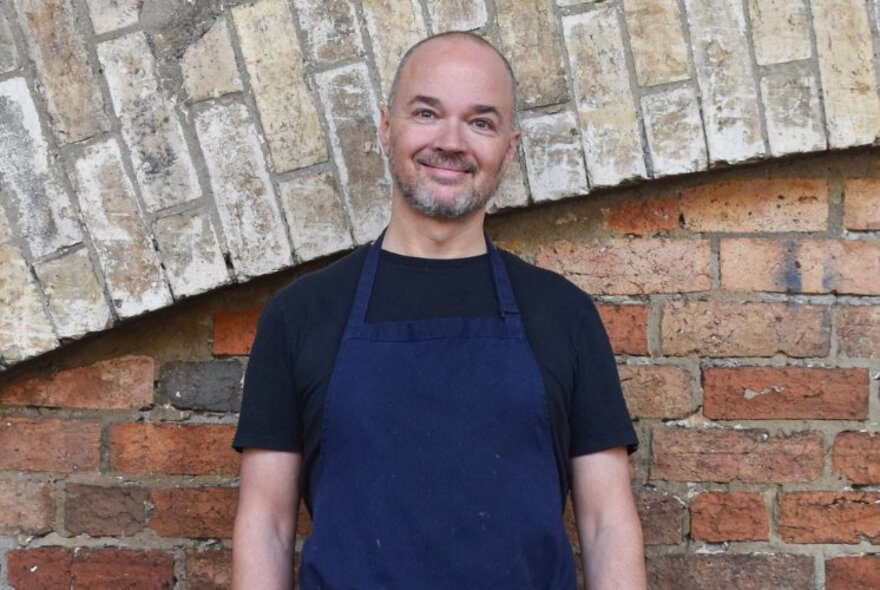 Chef Tobie Puttock, a smiling bald middle-aged man wearing a blue apron and standing in front of a brick wall.