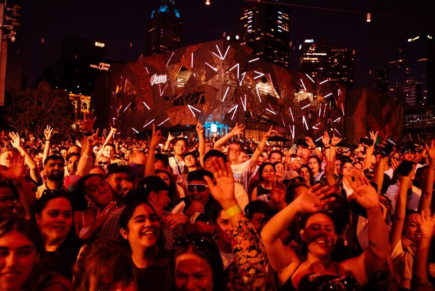 A crowd of people enjoying themselves at a music concert at Fed Square, nighttime.