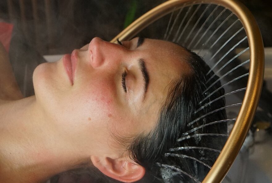 The relaxed face of a person lying back in a basin under a sprinkler system of water that is falling gently on her hair and scalp. 