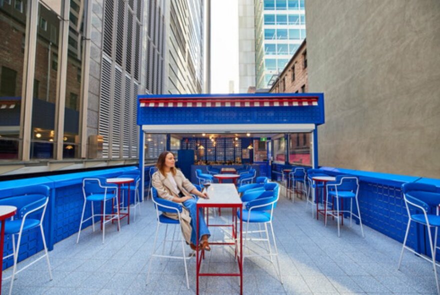 Woman in beige coat sitting at an outdoor table on blue coloured rooftop bar looking at the skyline.