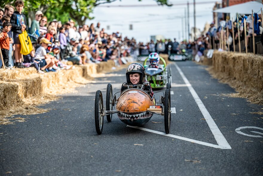 Two kids racing billy carts with a crowd watching on the sidelines.