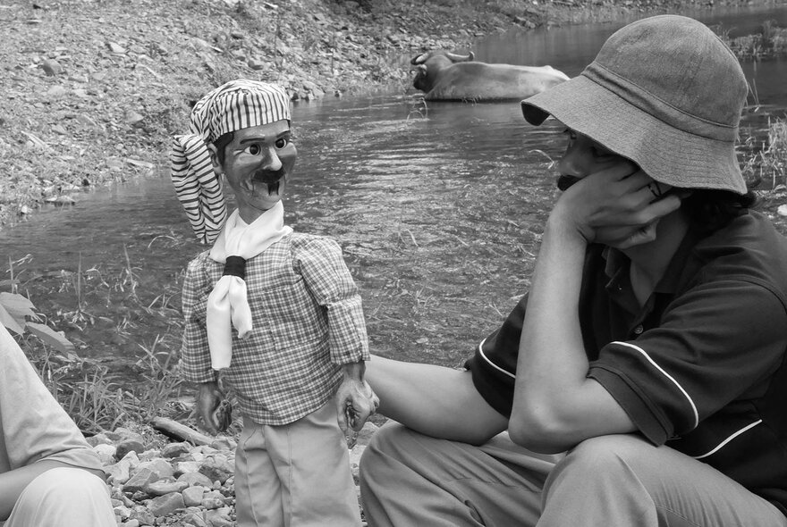 Black and white scene from the 2021 Philippine film History of Ha shows a person wearing a hat squatting near a river and holding a large puppet.