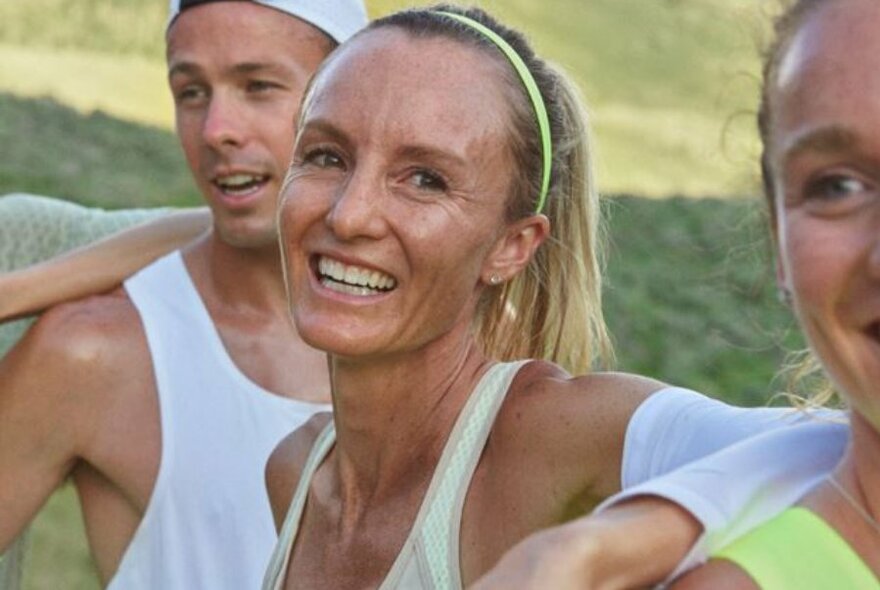 Woman with high ponytail and thin headband, wearing sporty tank top, sweaty and smiling, with two other people at side.