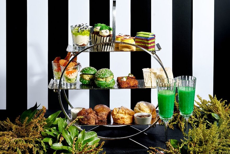 A circular three-tier cake stand with green Beetlejuice themed desserts surrounded by green foliage and a black and white stripe backdrop.