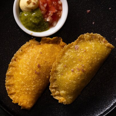 Two golden samosas on a black plate with a small white bowl of dipping sauces.