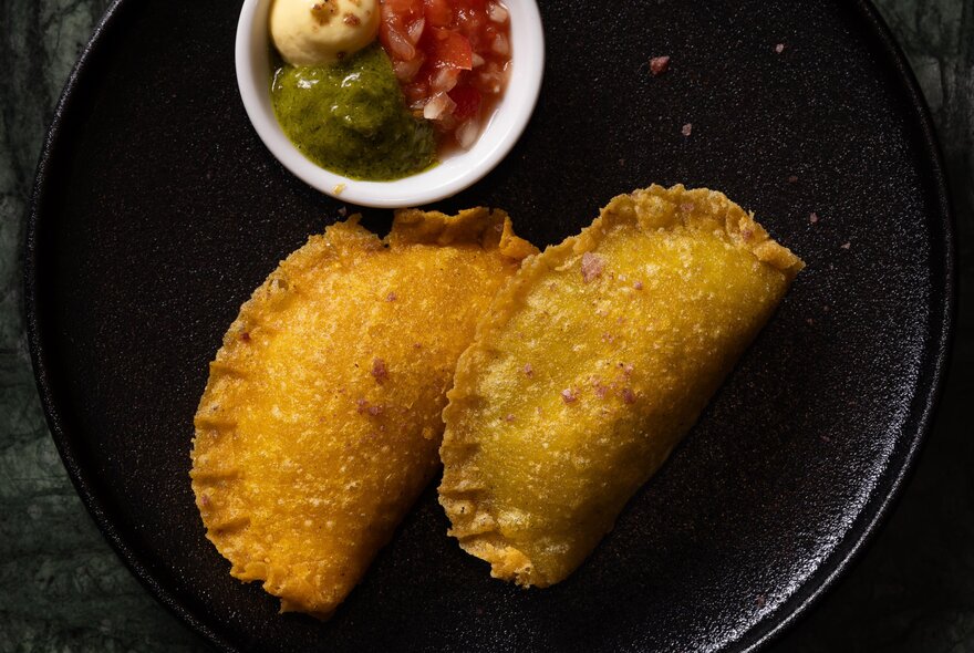 Two golden samosas on a black plate with a small white bowl of dipping sauces.