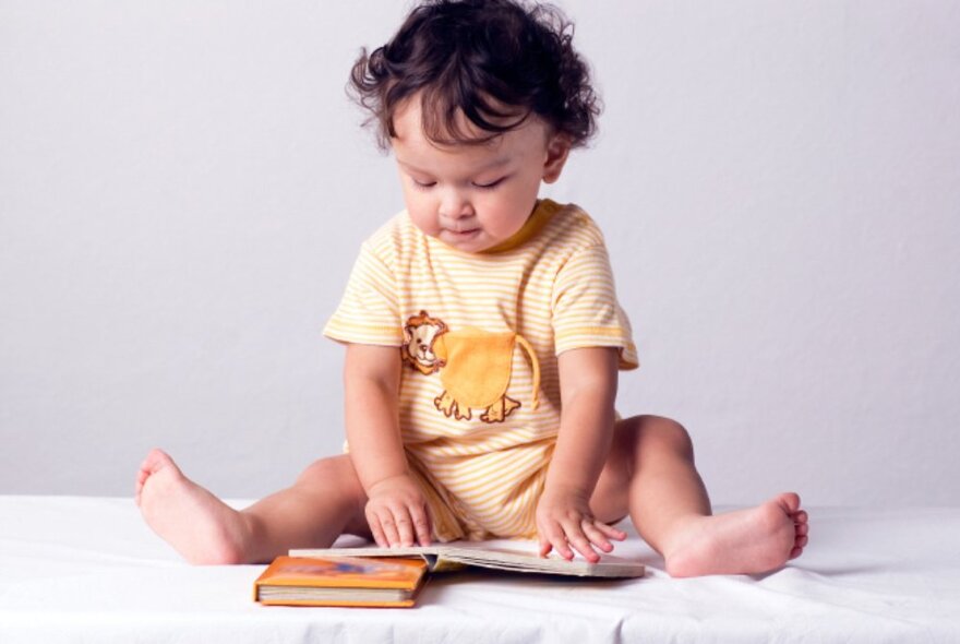 Toddler seated on a covered table reading books, with legs splayed.