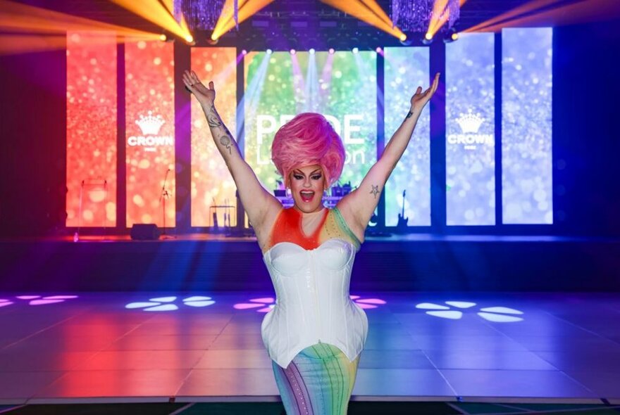 A drag queen with big pink hair and a while corset with hands in the air in a theatre setting. 