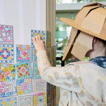 A child wearing a cardboard gladiator helmet adhering a mosaic-inspired square tile drawing onto a wall with other such designs, as part of an interactive workshop at the Melbourne Museum.
