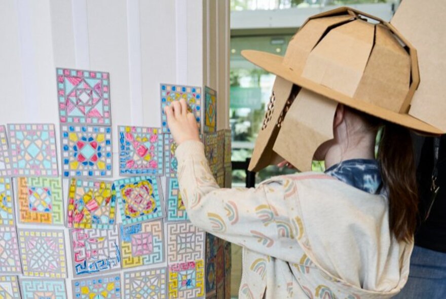 A child wearing a cardboard gladiator helmet adhering a mosaic-inspired square tile drawing onto a wall with other such designs, as part of an interactive workshop at the Melbourne Museum.