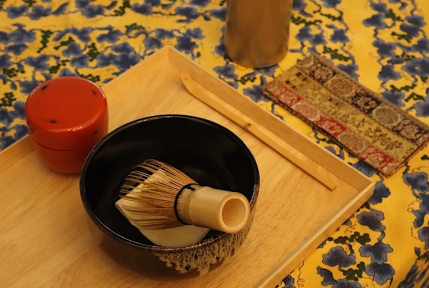 A traditional Japanese bowl with matcha whisk on a wooden tray, resting on a patterned tablecloth.