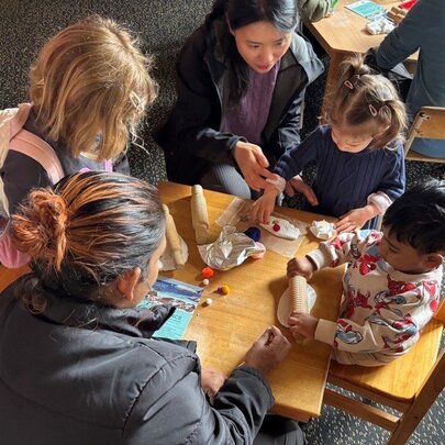 Kids sitting at a small table and chairs set with two adults helping them do craft. 