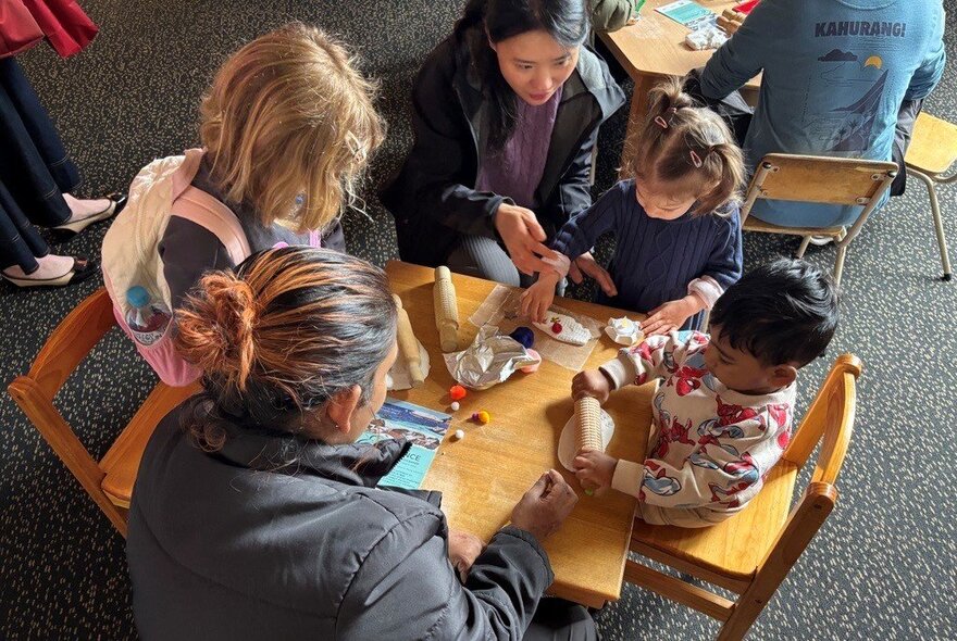 Kids sitting at a small table and chairs set with two adults helping them do craft. 