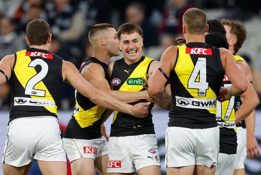 A huddle of Richmond AFL players celebrating on the field after having scored, with blurred fans in the stadium in the background.