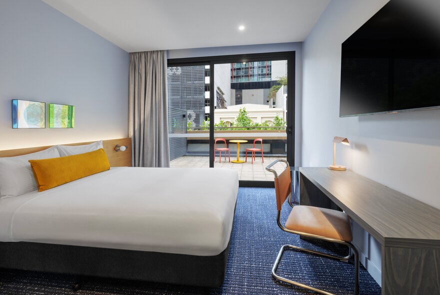 A clean hotel room with a bed, desk and chair, TV and an outdoor balcony with seating.