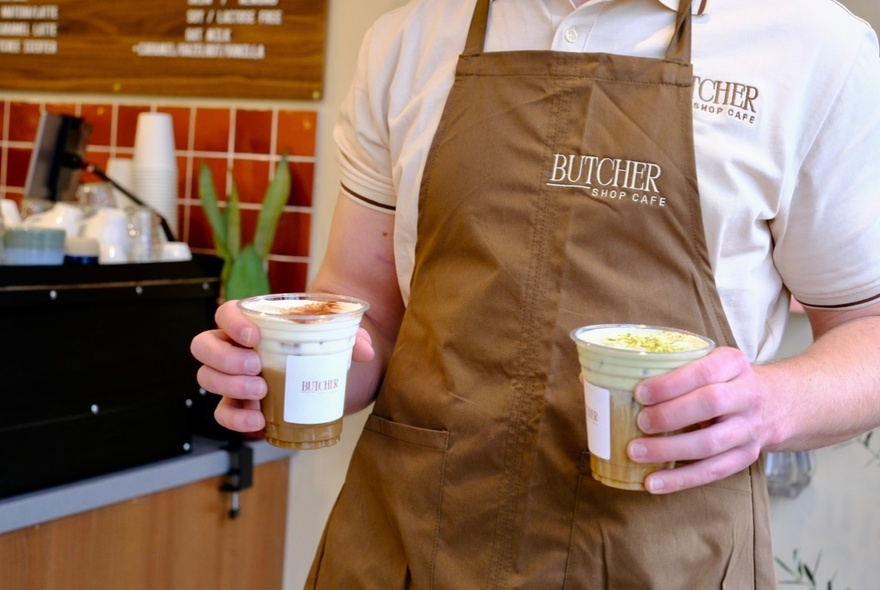 A person wearing a brown Butcher Shop Cafe branded apron, holding two takeaway cups.