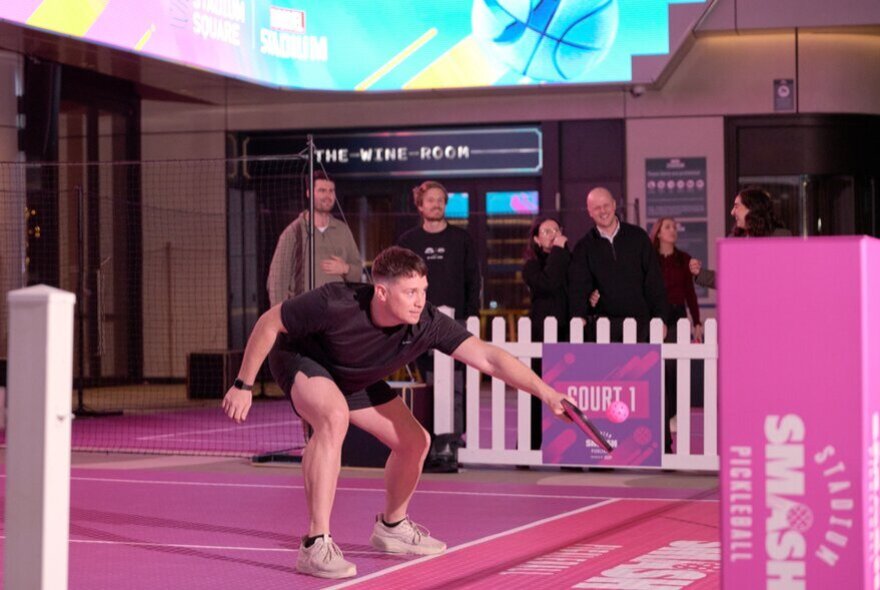 A man playing pickleball on a pickleball court inside Marvel Stadium, with people looking on from behind a low white picket fence.