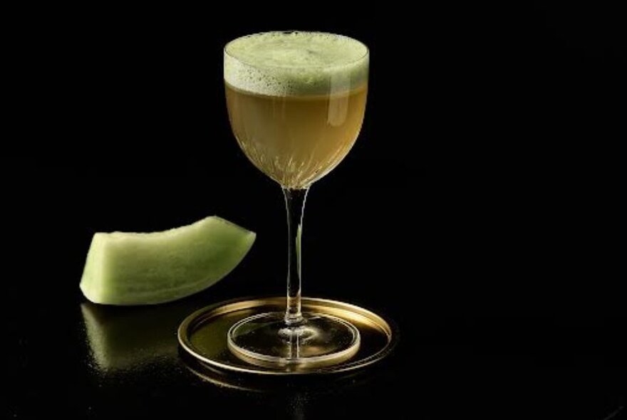 A green cocktail with a frothy green top, next to a slice of green melon.