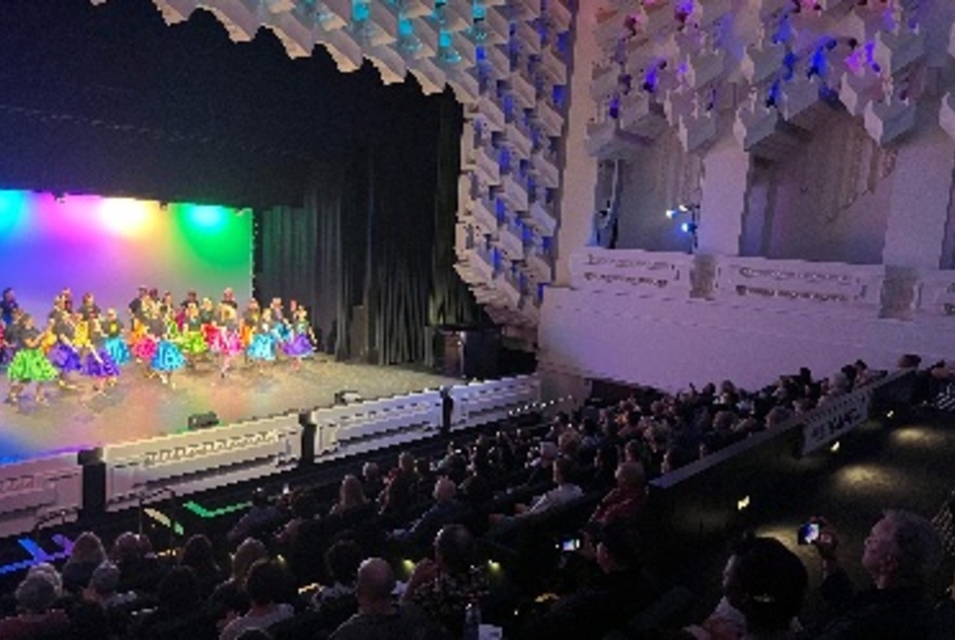 A colourful tap dancing troupe perfoming on stage at The Capitol, with a seated audience in front of them.