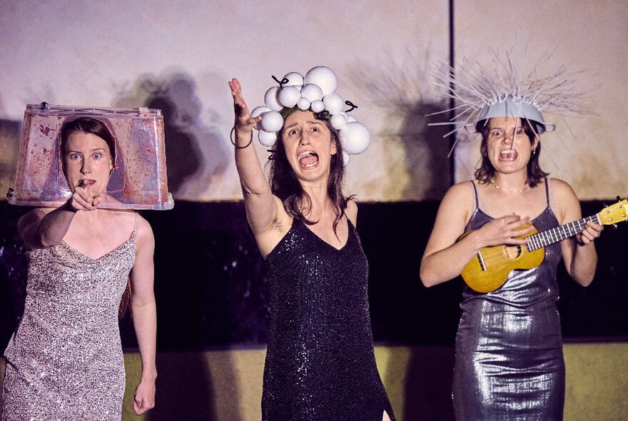 Three comedians standing with silly hats on their heads, one playing ukulele.