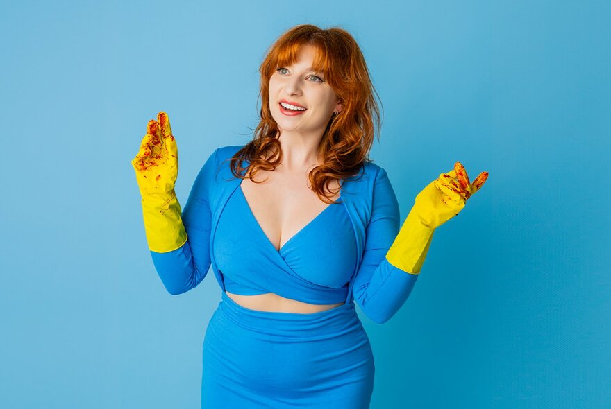 Performer Robyn Reynolds, a woman wearing an aqua coloured tight fitting dress, yellow rubbers gloves stained red at the fingertips, with shoulder length red hair and with a smile on her face.