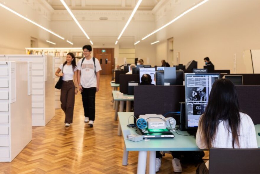 A room at the State Library Victoria, with people seated at desks looking at digitised newspapers on screens, and other people walking past rows of high drawer storage units.