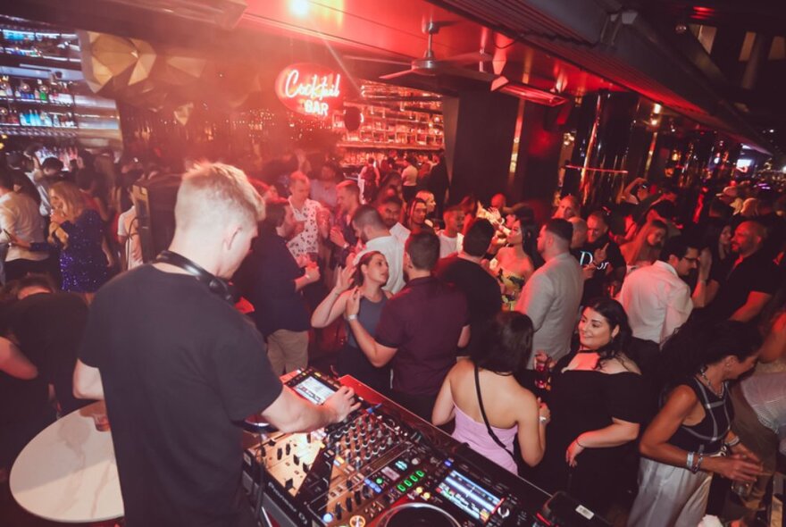 A DJ performing while people dance and mingle in a nightclub. 