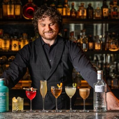 A smiling bartender looking down at a selection of cocktails in different shaped glasses on the bar that he stands behind.