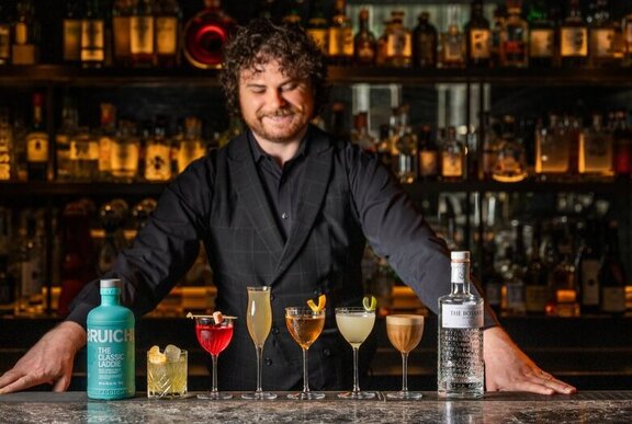 A smiling bartender looking down at a selection of cocktails in different shaped glasses on the bar that he stands behind.