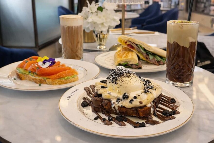 A brunch spread featuring an open-faced salmon toast, a loaded French toast or dessert plate with ice cream and chocolate drizzle, sandwiches, and iced drinks.