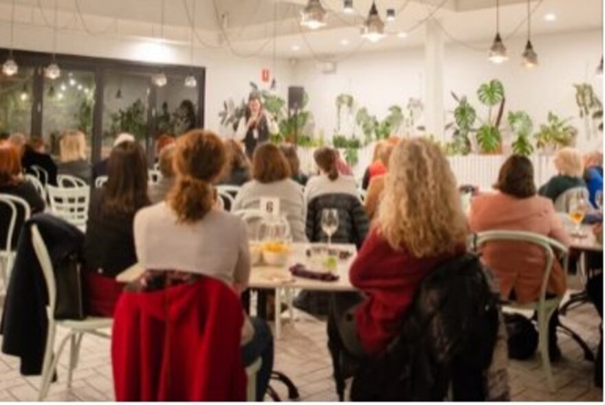 The back view of people sitting in a lit room listening to a speaker. 