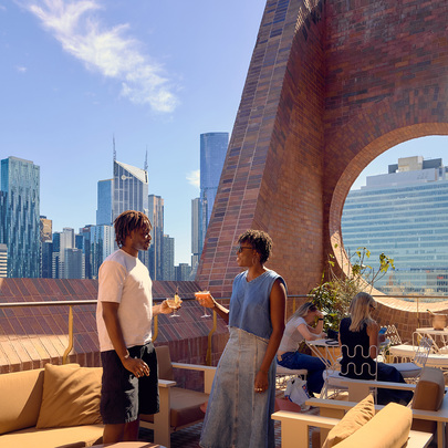 Two people drinking cocktails on an open air city rooftop bar with a brick wall with circular window.