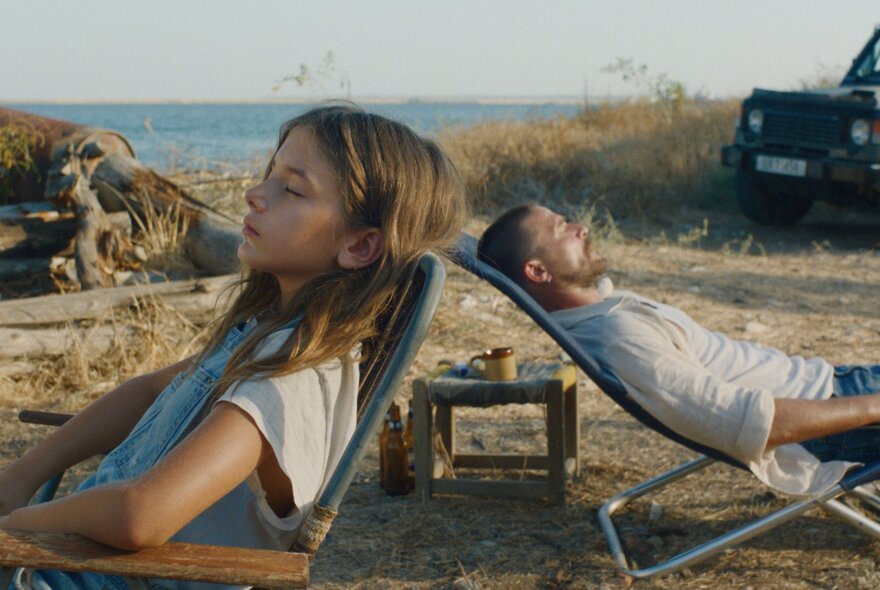 A still from a movie showing two people reclining in deck chairs in a grassy outdoor location, the sea and horizon visible in the distance.