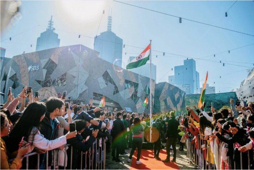 The Indian flag being raised at Fed Square, with a large crowd of onlookers cheering and celebrating in the open piazza.
