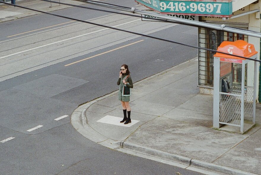 Person standing on a street corner near a phone box, wearing a short skirt and dark jacket with boots.