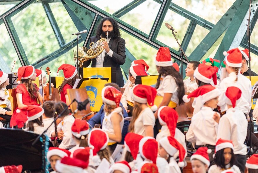 A choir of children wearing festive red Christmas hats looking at a musician playing a French horn.