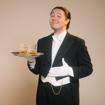 A person dressed in a formal tuxedo and white gloves holding a small silver tray with two whisky glasses on it.