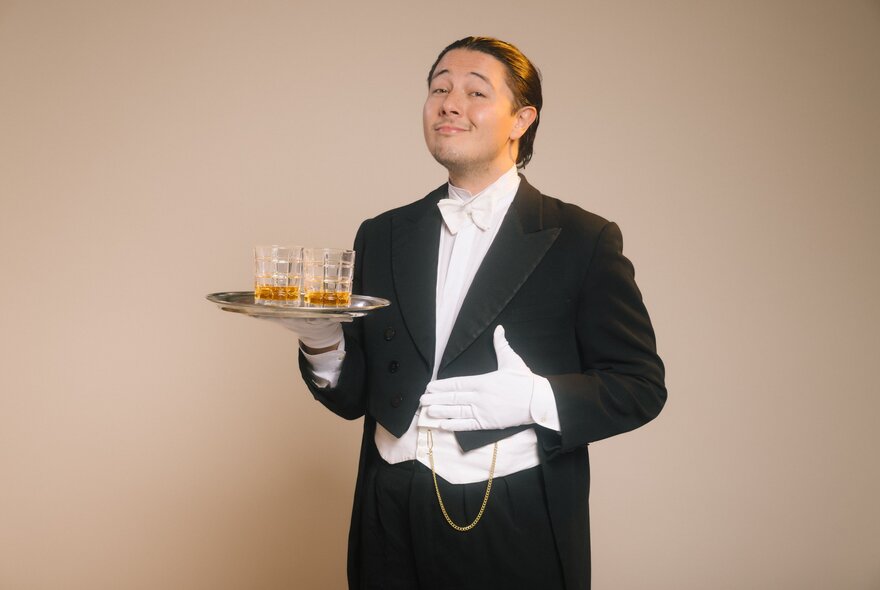 A person dressed in a formal tuxedo and white gloves holding a small silver tray with two whisky glasses on it.