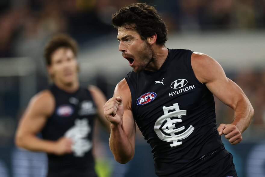 A Carlton AFL football player with pumped fists during a match, and another Carlton player behind him.