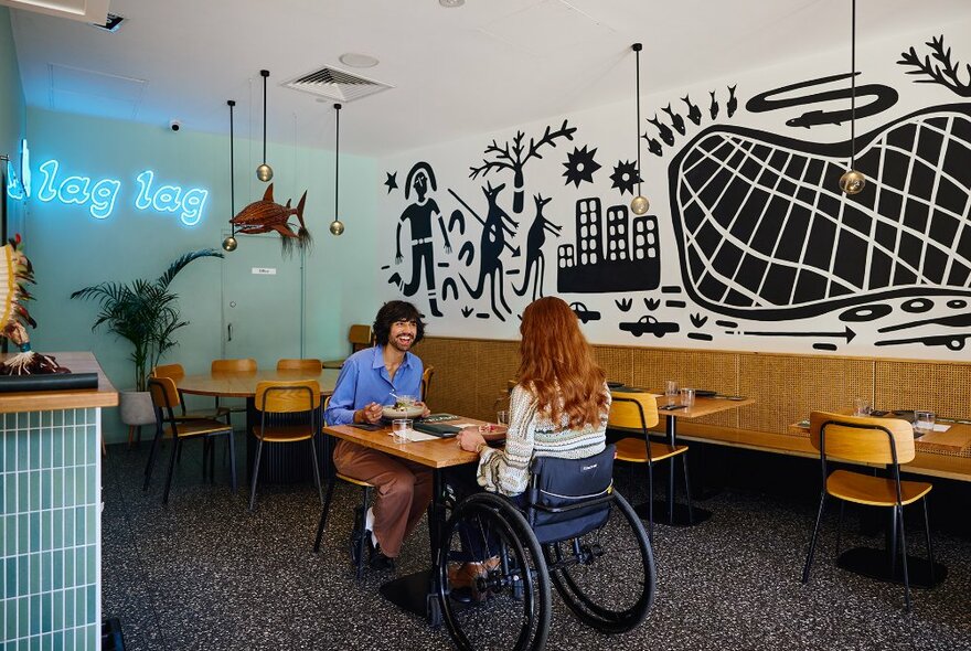 Two people sharing a meal in a restaurant with a black mural and blue neon sign. The woman is sitting in a wheelchair.