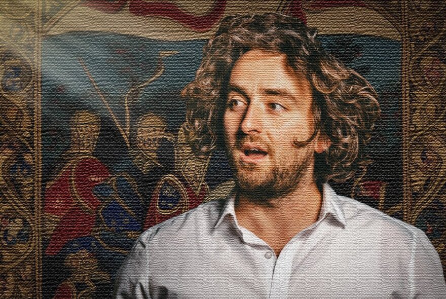 Comedian Joe Eidelson with long messy hair standing in front of a tapestry. 