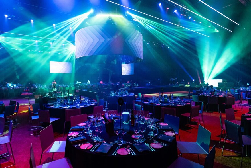 Round dining tables set for dinner and lit by turquoise strobe lighting in a large venue.