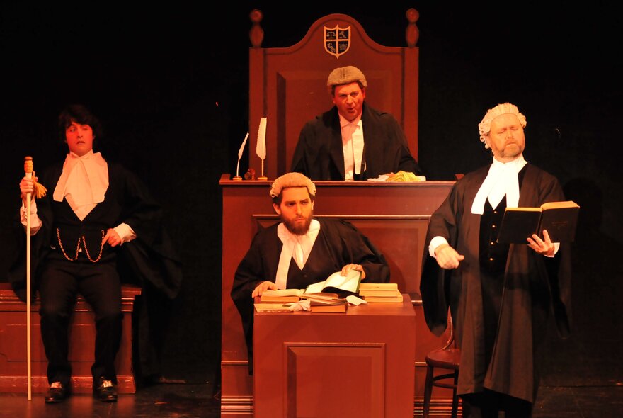 Actors in stage in a courtroom scene with lawyers in wigs and judge in an elevated seat. 